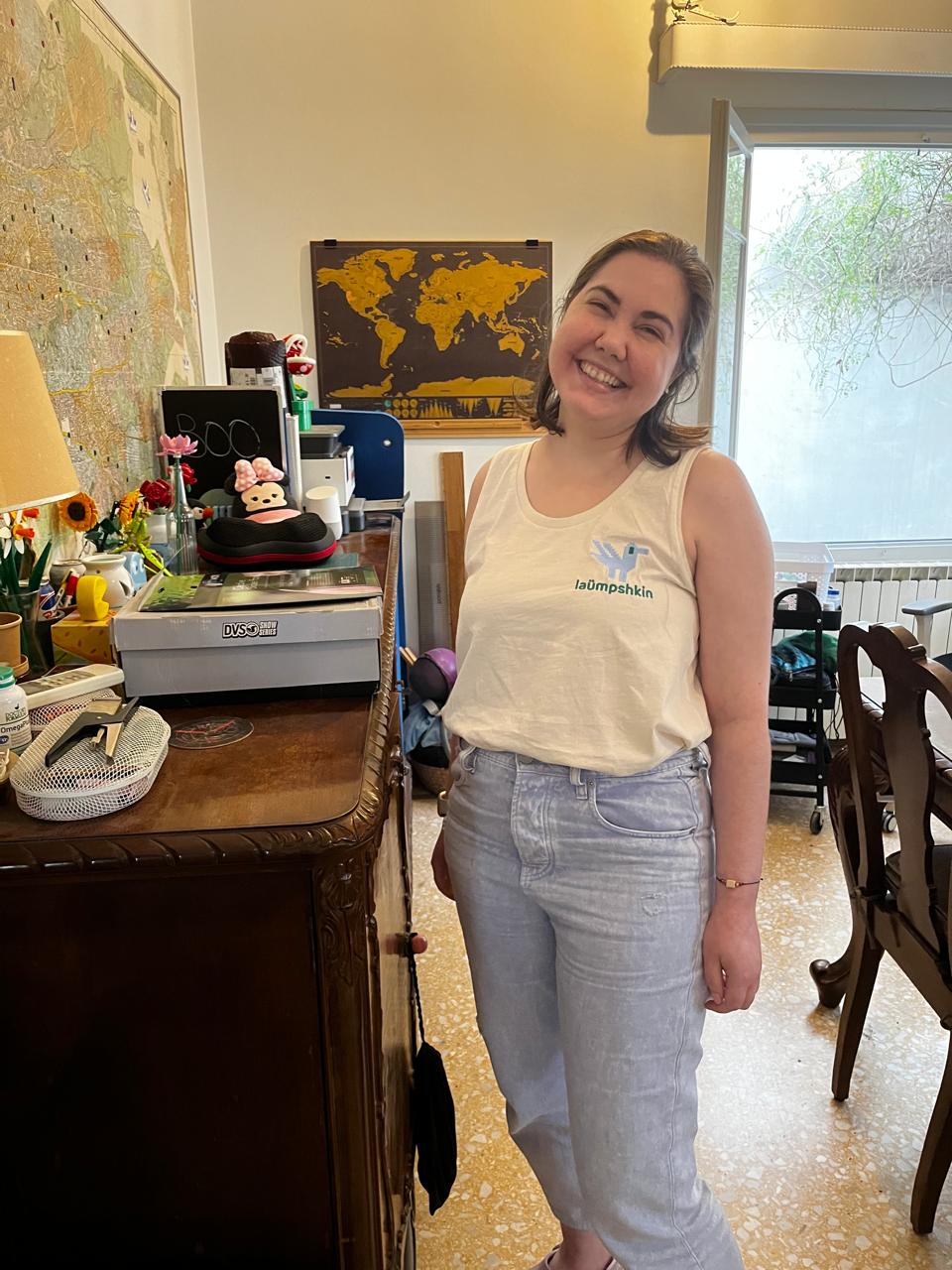 A disabled white woman standing in a messy office. She is wearing lavender jeans and crocs and an off-white tank top with a light blue duck embroidered on the top left. Under the duck is the word "laümpshkin" in bright green.