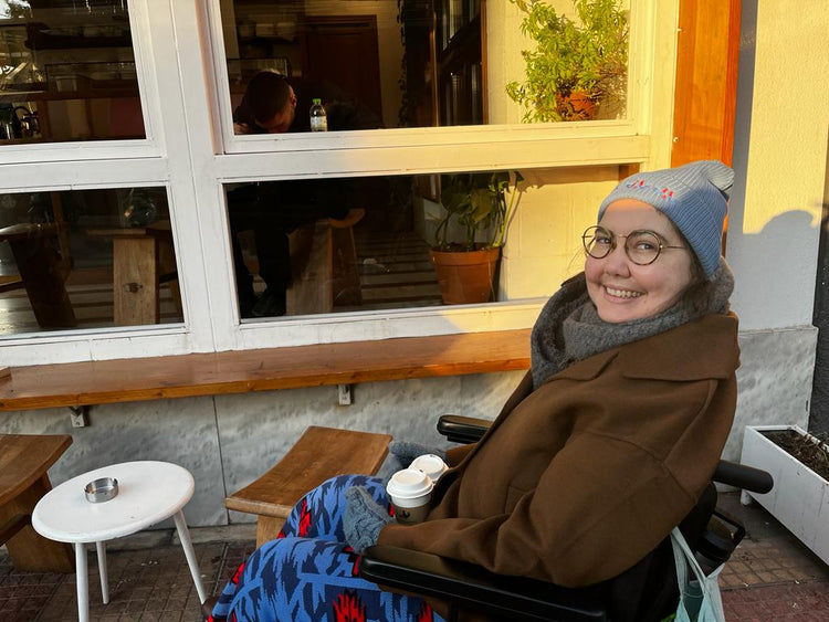 A woman with short brown hair and glasses sitting in a wheelchair in front of a cafe. She is wearing a light blue beanie with red and blue roosters embroidered on the front.