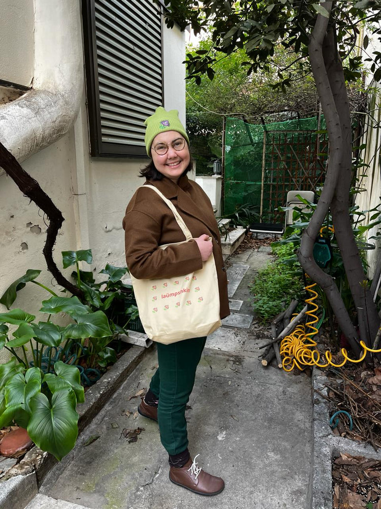 A woman with short brown hair and glasses standing in a garden. She is wearing a brown peacoat and holding a tan tote bag with the word "laümpshkin" printed on the front amongst rows of small peach flowers.