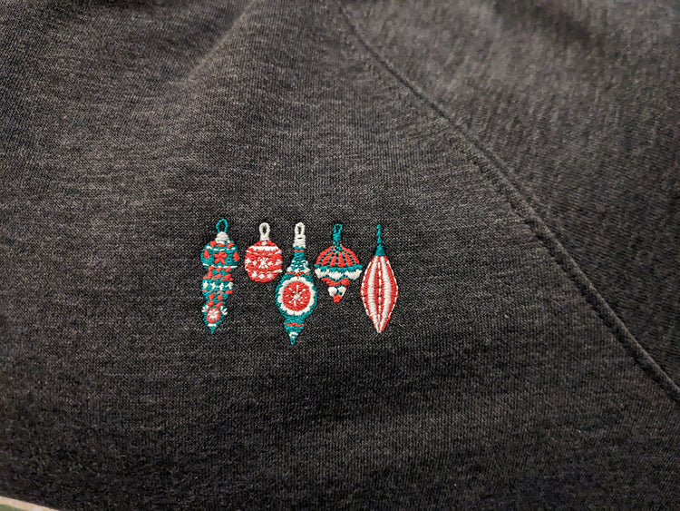 A close-up of the embroidery on a charcoal grey sweatshirt. It shows five Christmas ornaments in pink, green, and white.