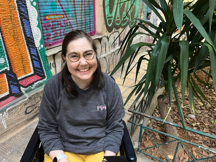 A woman with brown hair and glasses sits in her wheelchair in front of a wall of graffiti. She is wearing yellow pants and a charcoal grey sweatshirt with colorful ornaments embroidered on the top left.