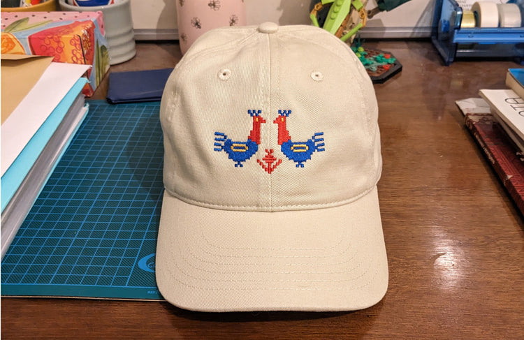 A tan baseball cap sitting on a messy desk. It has two large red and blue roosters with yellow details embroidered on the front.