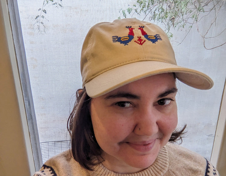 A woman with short brown hair looking at the camera and smiling. She is wearing a tan baseball cap with two large red and blue roosters with yellow details embroidered on the front.