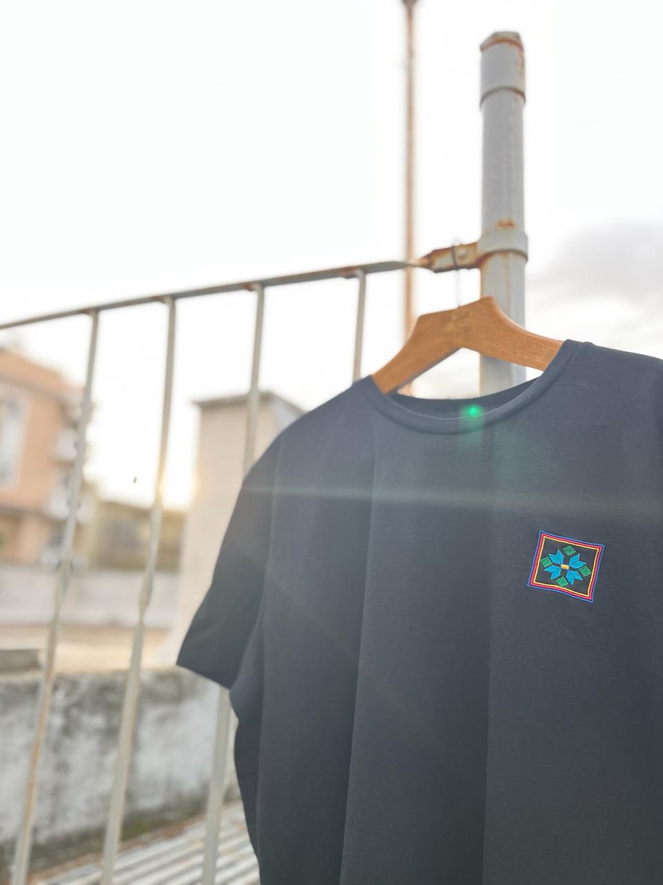 A 100% organic cotton black t-shirt is hanging on a railing with apartment buildings in the background.