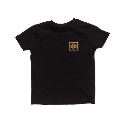 A black organic cotton t-shirt with an orange flower design embroidered on the top left. The flower is enclosed within three borders of yellow, pink, and blue.