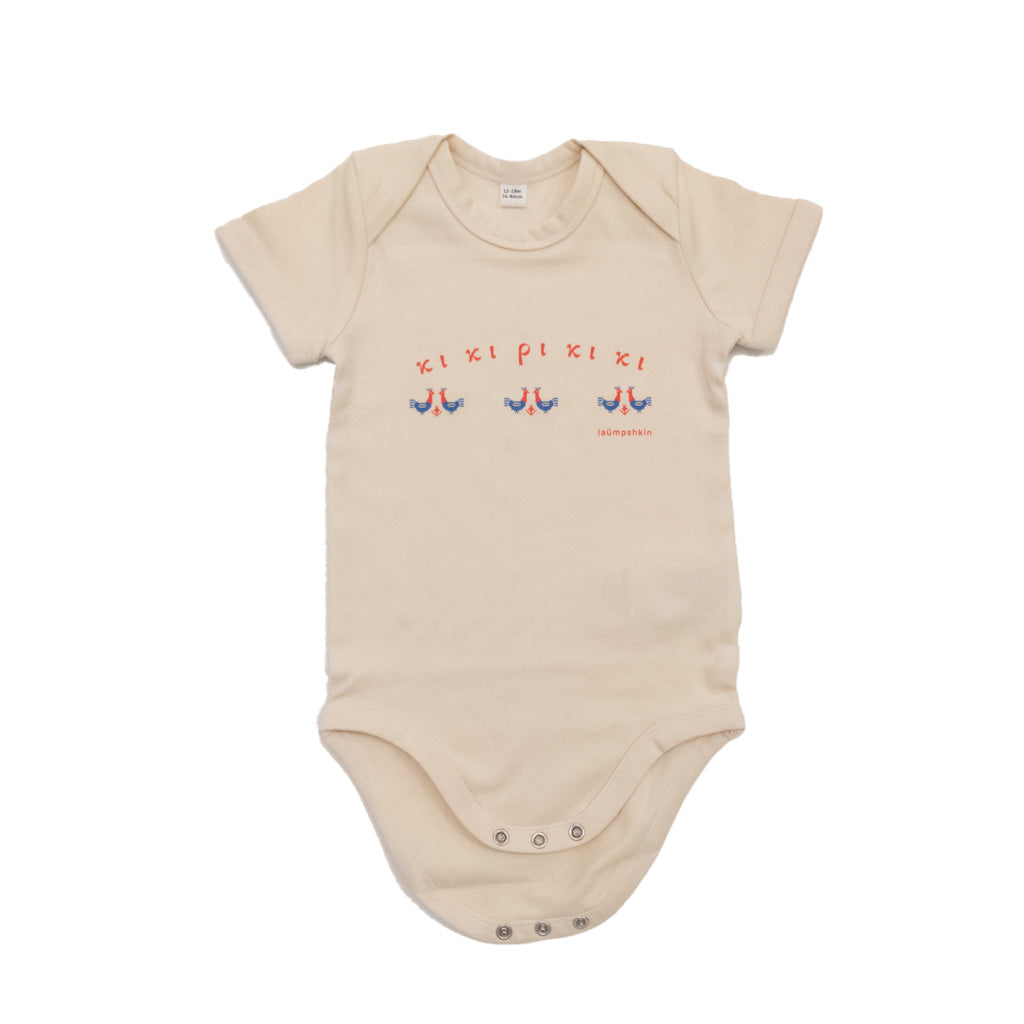 A short-sleeved beige baby bodysuit made of 100% organic cotton. On the front is a repeating design of red and blue roosters with the Greek words "κι κι ρι κι κι" above them.