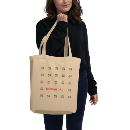 A woman holding a natural tan colored reusable shopping tote bag made of 100% organic cotton. Printed on the front is a 5x5 grid of small, intricate peach flowers, except for one on the second row, fourth column that is purple. On the second-to-last row, the middle three flowers are replaced by the word "laümpshkin".