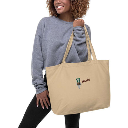 A woman holding a large beige festive holiday tote bag with a large embroidered design on the front. There is the head of a nutcracker wearing a green hat and a monocle and the word "Hark!". It is made of 100% organic cotton.