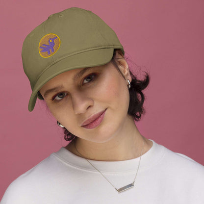 A woman wearing a khaki green baseball cap made of 100% organic cotton with an embroidered patch on the front. The patch is an orange circle with a yellow border with a purple retro duck inside.