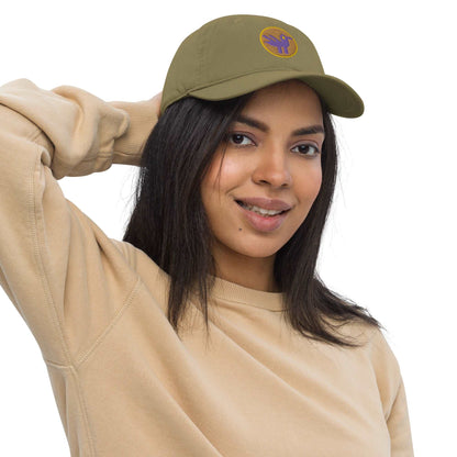 A woman wearing a khaki green baseball cap made of 100% organic cotton with an embroidered patch on the front. The patch is an orange circle with a yellow border with a purple retro duck inside.