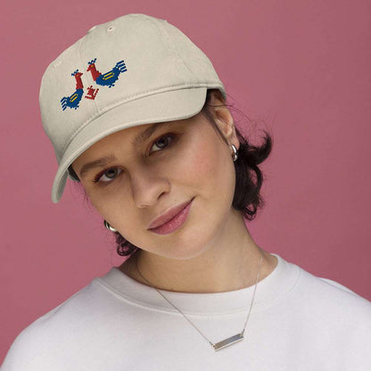 A woman wearing a baseball cap made of 100% organic cotton tilts her head and looks at the camera. The hat is a light tan color with two large red and blue roosters embroidered on the front.