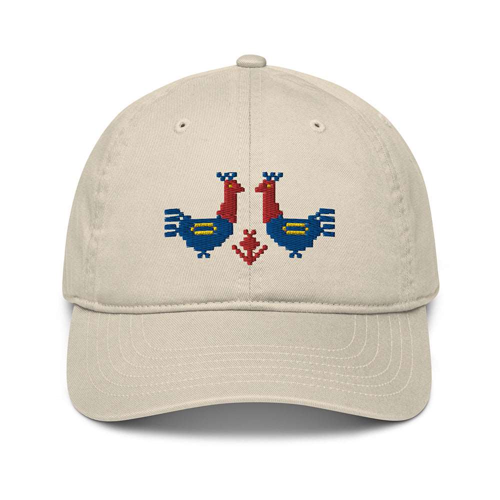 A light tan baseball cap made of 100% organic cotton with two large red and blue roosters embroidered on the front.