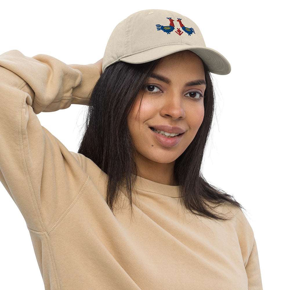 A woman wearing a baseball cap made of 100% organic cotton looking at the camera. The hat is a light tan color with two large red and blue roosters embroidered on the front.