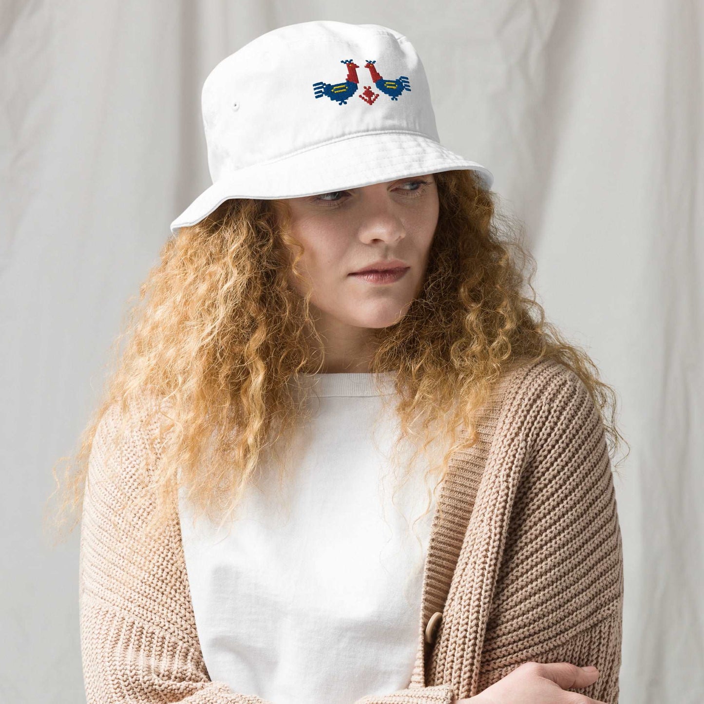 Roosters Organic bucket hat, a woman wearing a white bucket hat with a pair of large red and blue roosters with yellow details embroidered on the front.