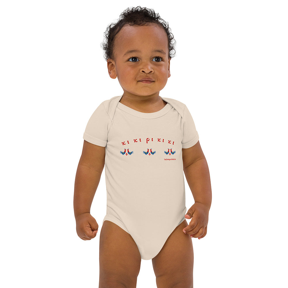 A standing baby wearing a short-sleeved beige baby bodysuit made of 100% organic cotton. On the front is a repeating design of red and blue roosters with the Greek words "κι κι ρι κι κι" above them.