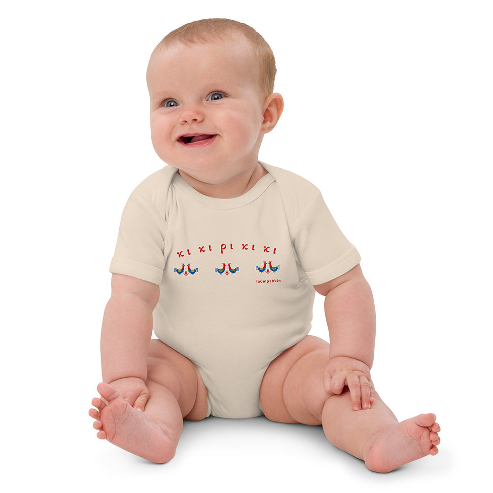 A seated baby wearing a short-sleeved beige baby bodysuit made of 100% organic cotton. On the front is a repeating design of red and blue roosters with the Greek words "κι κι ρι κι κι" above them.