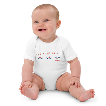 A seated baby wearing a short-sleeved white baby bodysuit made of 100% organic cotton. On the front is a repeating design of red and blue roosters with the Greek words "κι κι ρι κι κι" above them.