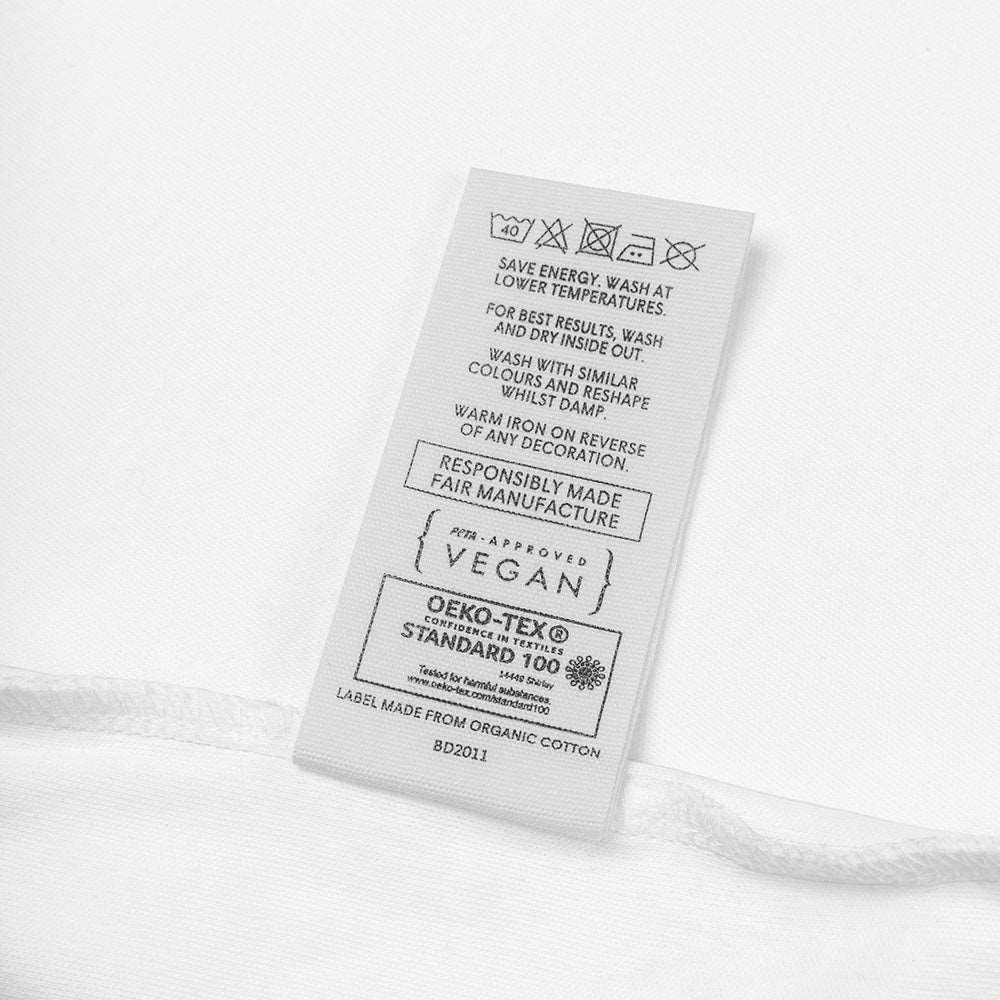 Detail of the tag on a white baby bodysuit made of organic cotton. It shows the laundry care instructions, that it is Peta-Approved vegan, and the Oeko-Tek label.