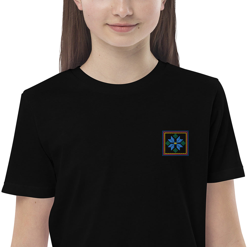 A close-up of a girl wearing a black organic cotton t-shirt with a blue flower design embroidered on the top left. The flower is enclosed within three borders of yellow, pink, and blue.