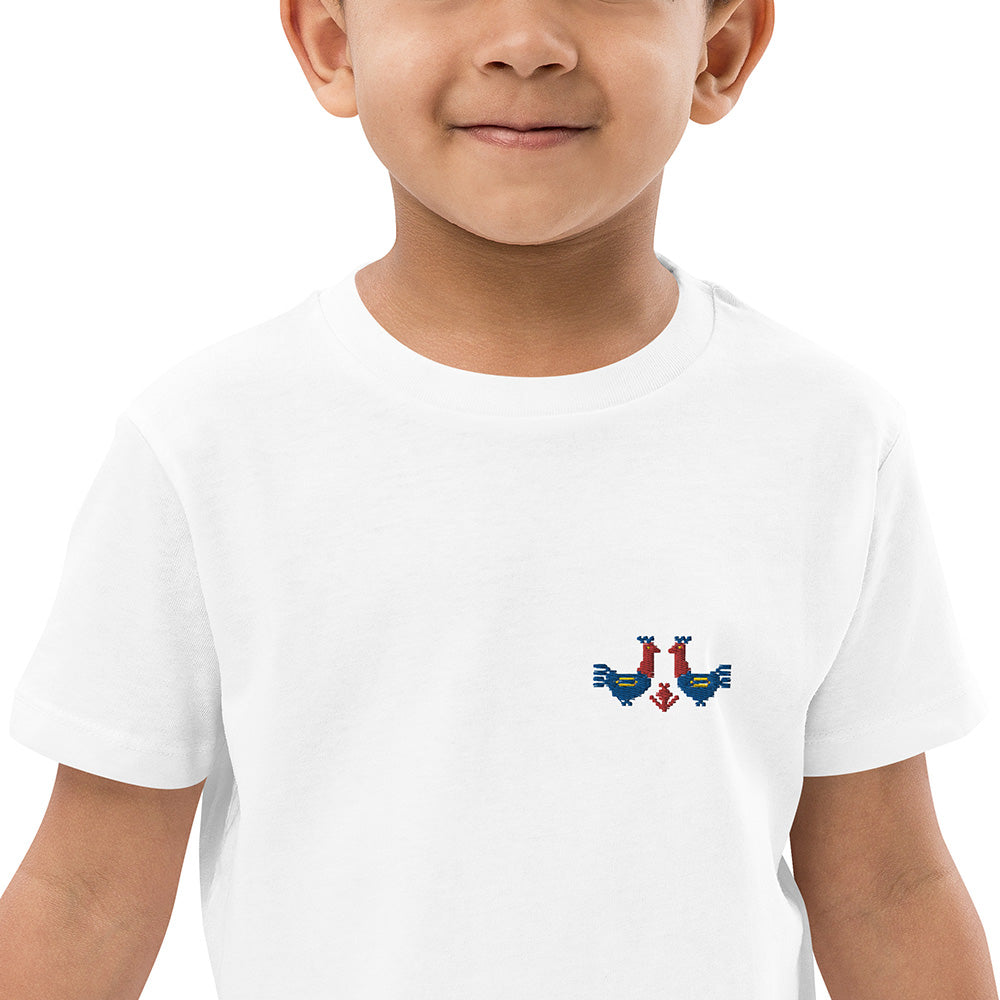 A close-up of a boy wearing a white t-shirt made of 100% organic cotton with a pair of red and blue roosters embroidered on the top left.