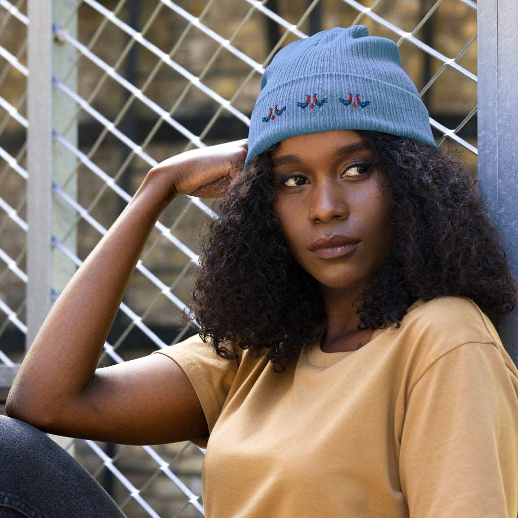 A woman wearing a light blue 100% organic cotton ribbed beanie with 3 pairs of red and blue roosters embroidered on the front.