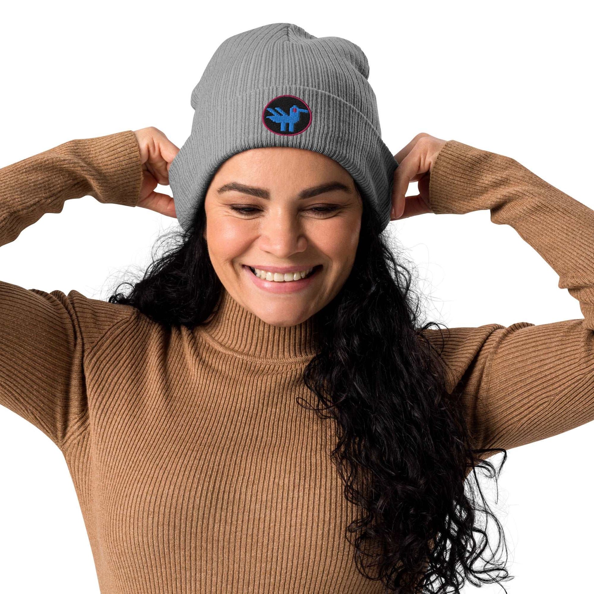 A woman wearing a grey beanie made of 100% organic cotton with a patch embroidered on the front. The patch is a black circle with a pink border with a blue retro style duck inside.