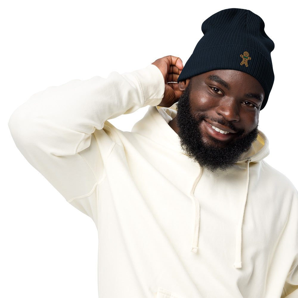 A man wearing a navy blue ribbed festive holiday beanie with a gingerbread man embroidered on the front. The beanie is made of 100% organic cotton.