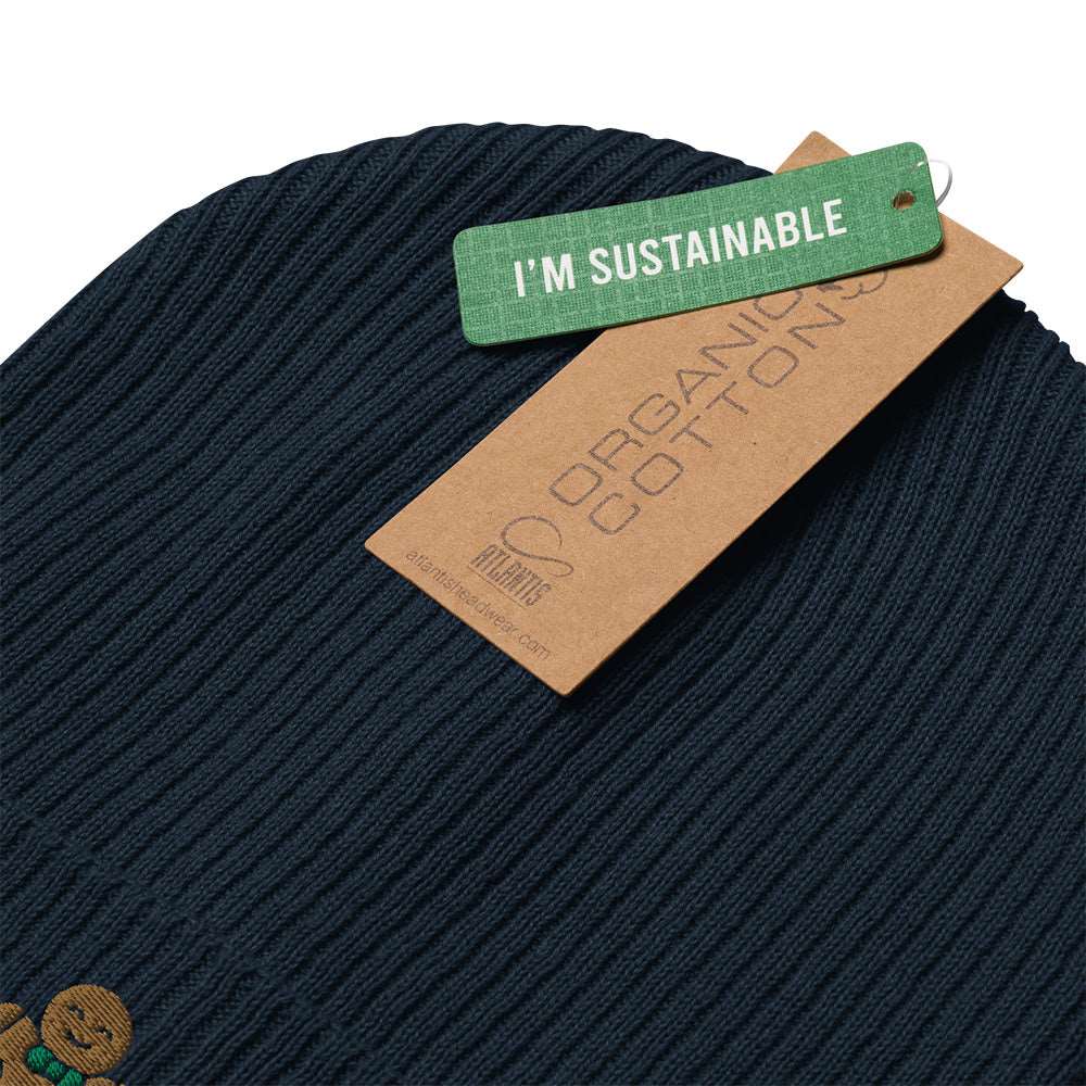 Detail of the tags on a navy blue ribbed holiday beanie with a gingerbread man embroidered on the front. The tags say "I'm Sustainable" and "Organic Cotton". It is made of 100% organic cotton.