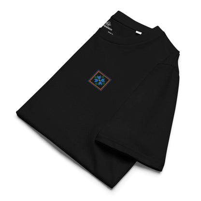 A black organic cotton t-shirt with a blue flower design embroidered on the top left. The flower is enclosed within three borders of yellow, pink, and blue. It is folded and shows the top left embroidered detail and the left sleeve.