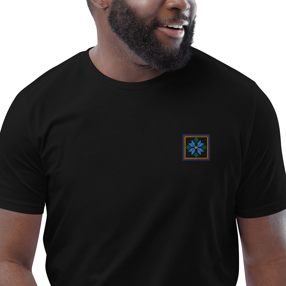 A close-up of a man wearing a black organic cotton t-shirt with a blue flower design embroidered on the top left. The flower is enclosed within three borders of yellow, pink, and blue.