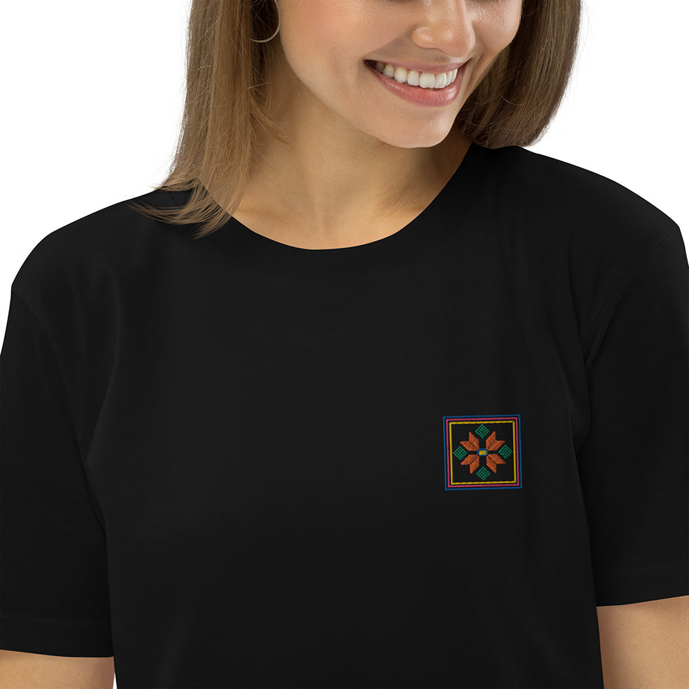 A close-up of a woman wearing a black organic cotton t-shirt with an orange flower design embroidered on the top left. The flower is enclosed within three borders of yellow, pink, and blue.