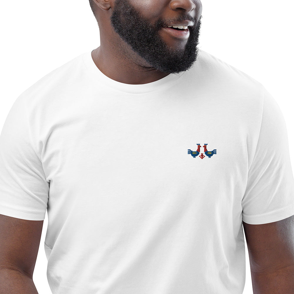 A close-up of a man wearing a white t-shirt made of 100% organic cotton with a pair of red and blue roosters embroidered on the top left.