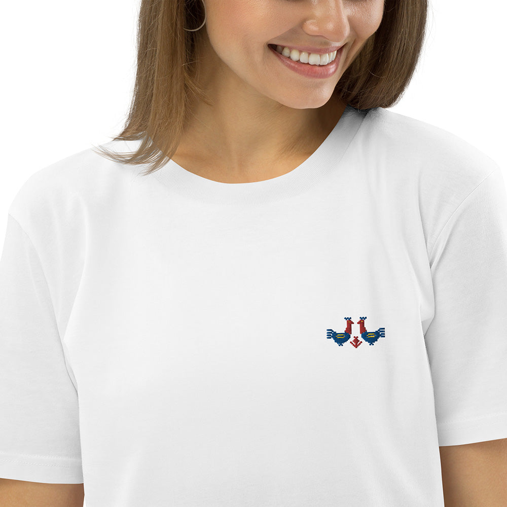 A close-up of a woman wearing a white t-shirt made of 100% organic cotton with a pair of red and blue roosters embroidered on the top left.