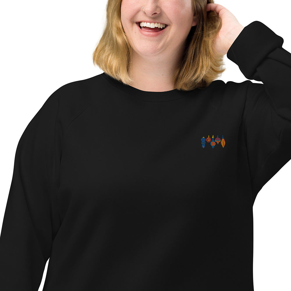 A closeup of a woman wearing a 100% organic cotton black sweatshirt with four small Christmas ornaments embroidered on the top left. The ornaments have varying shapes and patterns and are colored blue, red, and yellow.